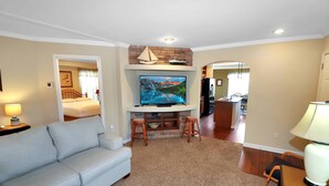 A living room with a sofa, TV, and a fireplace. There is a kitchen in the background. - My Island Escape | Luxury New Home, Gourmet Kitchen, Soaking Tub, Deck Retreat Near North Main (Chincoteague Island)