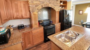 A kitchen with wooden cabinets, a granite countertop, a black stove, a stainless steel sink, and a stone arch above the stove. - My Island Escape | Luxury New Home, Gourmet Kitchen, Soaking Tub, Deck Retreat Near North Main (Chincoteague Island)
