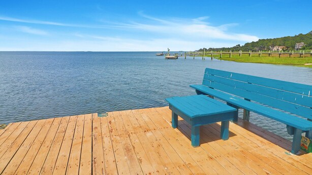 A wooden pier with benches overlooking a calm body of water, a distant pier, and a grassy area with houses in the background. - My Island Escape | Luxury New Home, Gourmet Kitchen, Soaking Tub, Deck Retreat Near North Main (Chincoteague Island)
