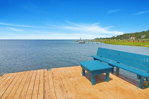 A wooden pier with benches overlooking a calm body of water, a distant pier, and a grassy area with houses in the background.