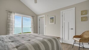A bedroom with a large bed, a view of the sea, a door, and a chair. - Poole's Cove | Waterfront Main Street Retreat Fully Renovated w/ Rooftop Lounge & Artistic Charm (Chincoteague Island)