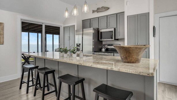 A modern kitchen with a large island, bar stools, and a view to the outside. - Poole's Cove | Waterfront Main Street Retreat Fully Renovated w/ Rooftop Lounge & Artistic Charm (Chincoteague Island)