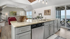 A modern kitchen with a large island, a dishwasher, and a dining area with a view. - Poole's Cove | Waterfront Main Street Retreat Fully Renovated w/ Rooftop Lounge & Artistic Charm (Chincoteague Island)
