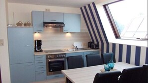 A compact kitchen with light blue cabinets, a stove, and a sink. There is a dining table with chairs and a blue vase on it. A window with a striped curtain is visible.