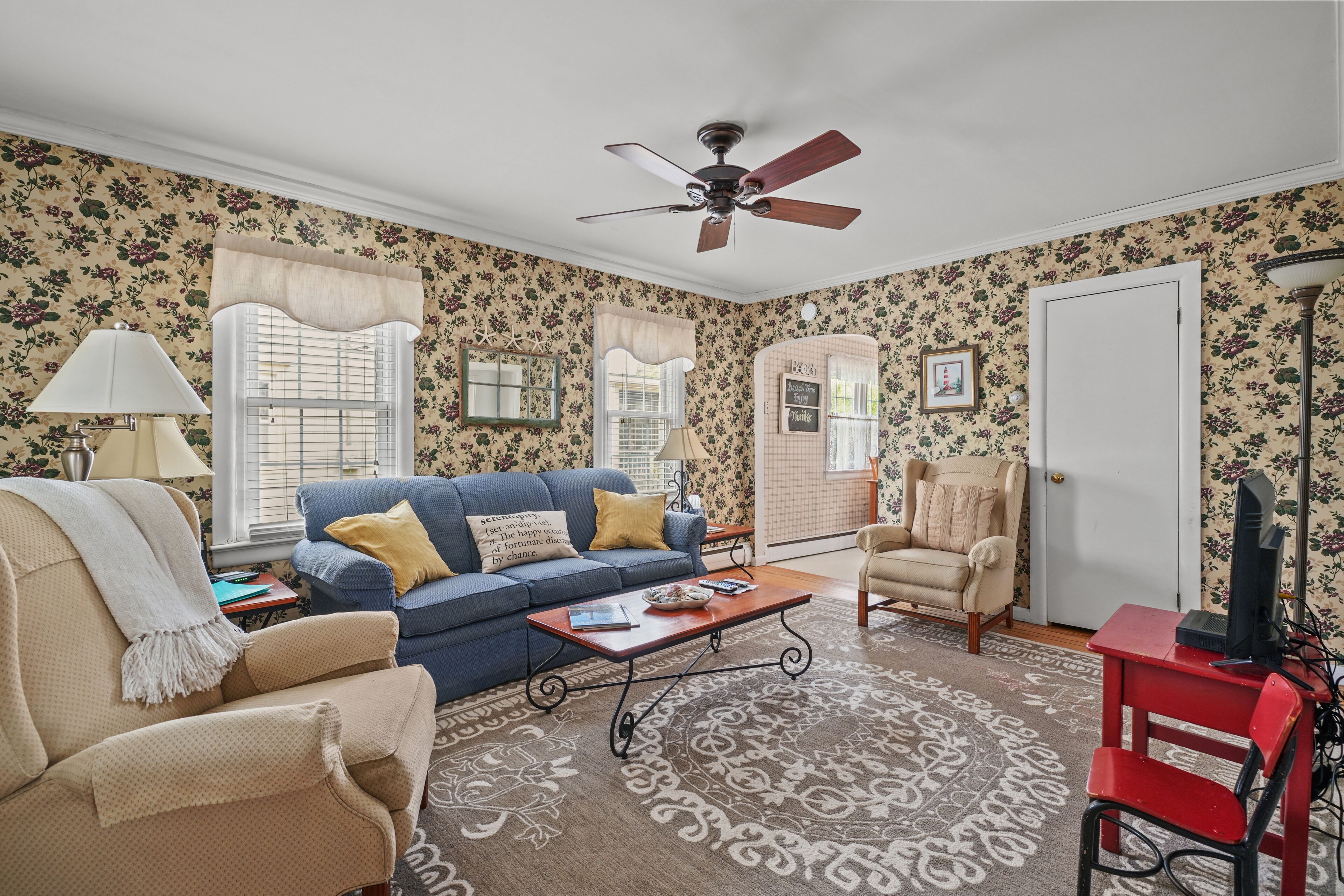 A living room with floral wallpaper, a ceiling fan, a blue sofa, a beige armchair, a red chair, and a television.