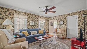 A living room with floral wallpaper, a ceiling fan, a blue sofa, a beige armchair, a red chair, and a television. - Serendipity | Classic Chincoteague Home Walk to Main Street w/ Screened Porch & First-Floor Bedroom (Chincoteague Island)