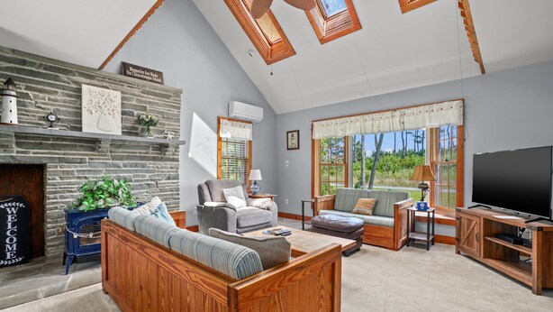 A spacious living room with a stone fireplace, wooden furniture, and large windows offering a view of greenery. - Shady Side | Piney Island Home Near Beach w/ Community Water Access, Large Porch, Grill & Loft (Chincoteague Island)