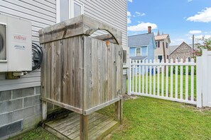 A wooden structure with a metal roof, possibly a shed or storage unit, adjacent to a house with a white fence and a Fujitsu air conditioning unit.