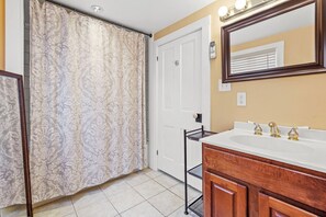 Un baño con cortina de ducha estampada, una puerta blanca, tocador con espejo y lavabo con tres grifos.