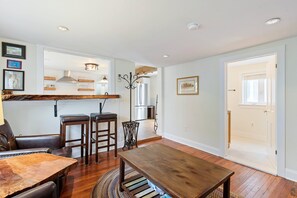 Uma sala de estar espaçosa com uma mesa de madeira e dois bancos altos, uma cozinha com um exaustor de aço inoxidável e uma porta que dá para outro quarto.