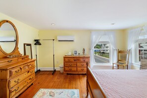 A bedroom with a wooden dresser, a bed with a pink quilt, a mirror, an air conditioning unit, and a view of the street through a window.