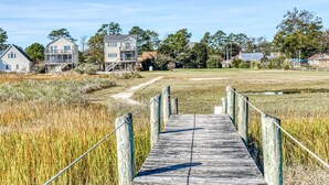 A wooden boardwalk leading to a residential area with houses and a grassy field. - Wildflower | Waterfront w/ Shared Pier & Floating Dock, Screened Porches + Balcony Views (Chincoteague Island)