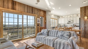 A spacious living area with a large window, a sofa, a coffee table, and a dining table. - Wildflower | Waterfront w/ Shared Pier & Floating Dock, Screened Porches + Balcony Views (Chincoteague Island)