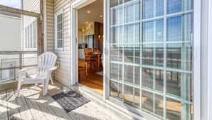 A covered deck with a white chair, a patterned rug, and a sliding glass door leading to a kitchen area with wooden cabinets and a dining table. - Wildflower | Waterfront w/ Shared Pier & Floating Dock, Screened Porches + Balcony Views (Chincoteague Island)