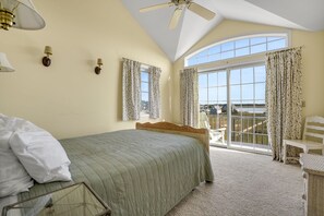 A bedroom with a bed, a ceiling fan, a window with curtains, and a balcony with a chair. - Wildflower | Waterfront w/ Shared Pier & Floating Dock, Screened Porches + Balcony Views (Chincoteague Island)