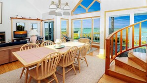 A spacious living area with a large dining table, comfortable seating, a television, and a scenic view of the ocean through large windows.