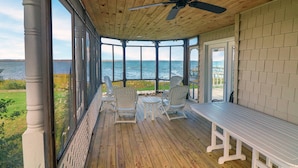 A covered porch with a view of the water, wooden flooring, white chairs, and a table.