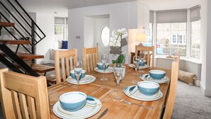 A dining area with a wooden table set for six, surrounded by wooden chairs. A staircase with black railings is visible to the left. - Bodwena (Moelfre)