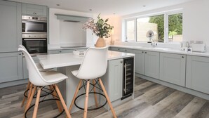 A modern kitchen with two white chairs, a built-in oven, a sink, and a window with a view of greenery. - Bodwena (Moelfre)