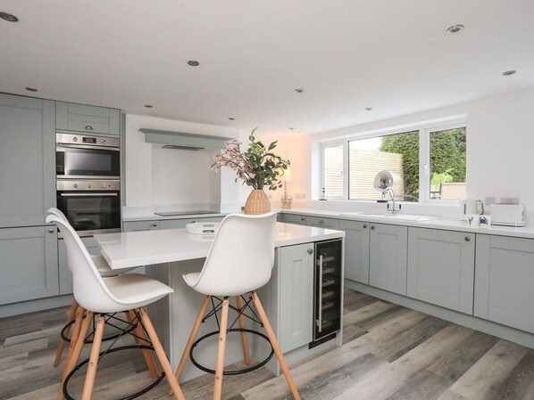 A modern kitchen with two white chairs, a built-in oven, a sink, and a window with a view of greenery.