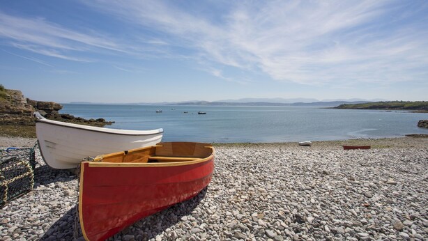A rocky beach with two boats, one white and one red, and calm water in the background. - Bodwena (Moelfre)