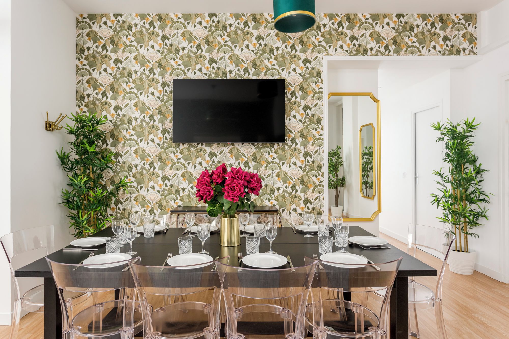 Un coin repas avec une table noire dressée pour six personnes, entourée de chaises transparentes. Un grand miroir et un téléviseur à écran plat sont fixés au mur.