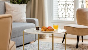 Een gezellige woonkamer met een grijze bank, een klein rond tafeltje met croissants, een kop koffie en een glas sinaasappelsap.
