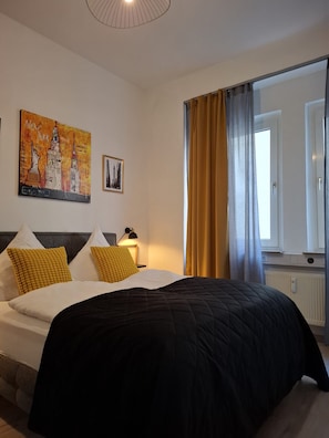 A hotel room with a bed, a painting of New York City, a nightstand, a lamp, and a window with curtains.