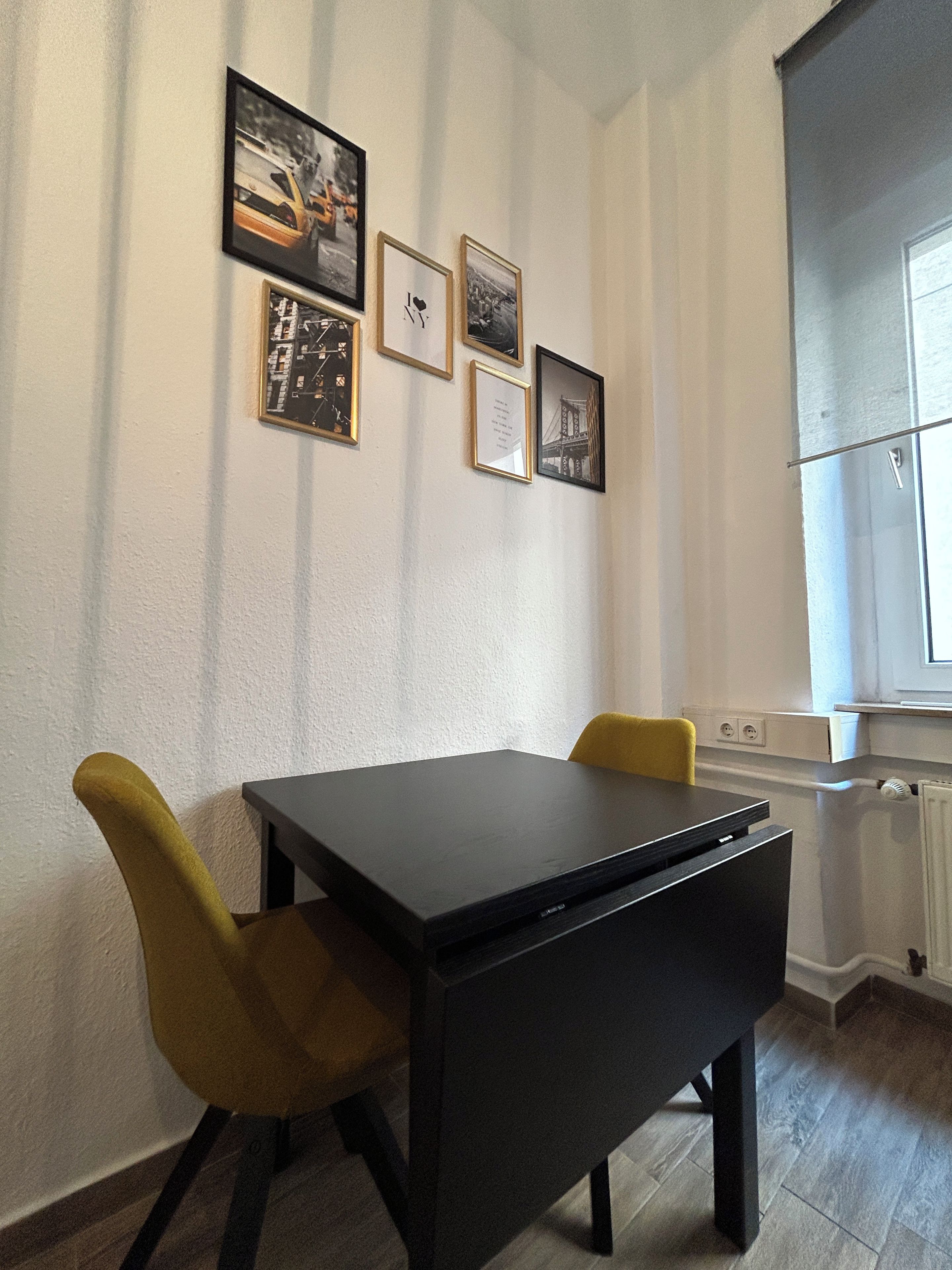 Ein Essbereich mit Tisch und Stühlen, ein Fenster mit Jalousien und gerahmte Bilder an der Wand.