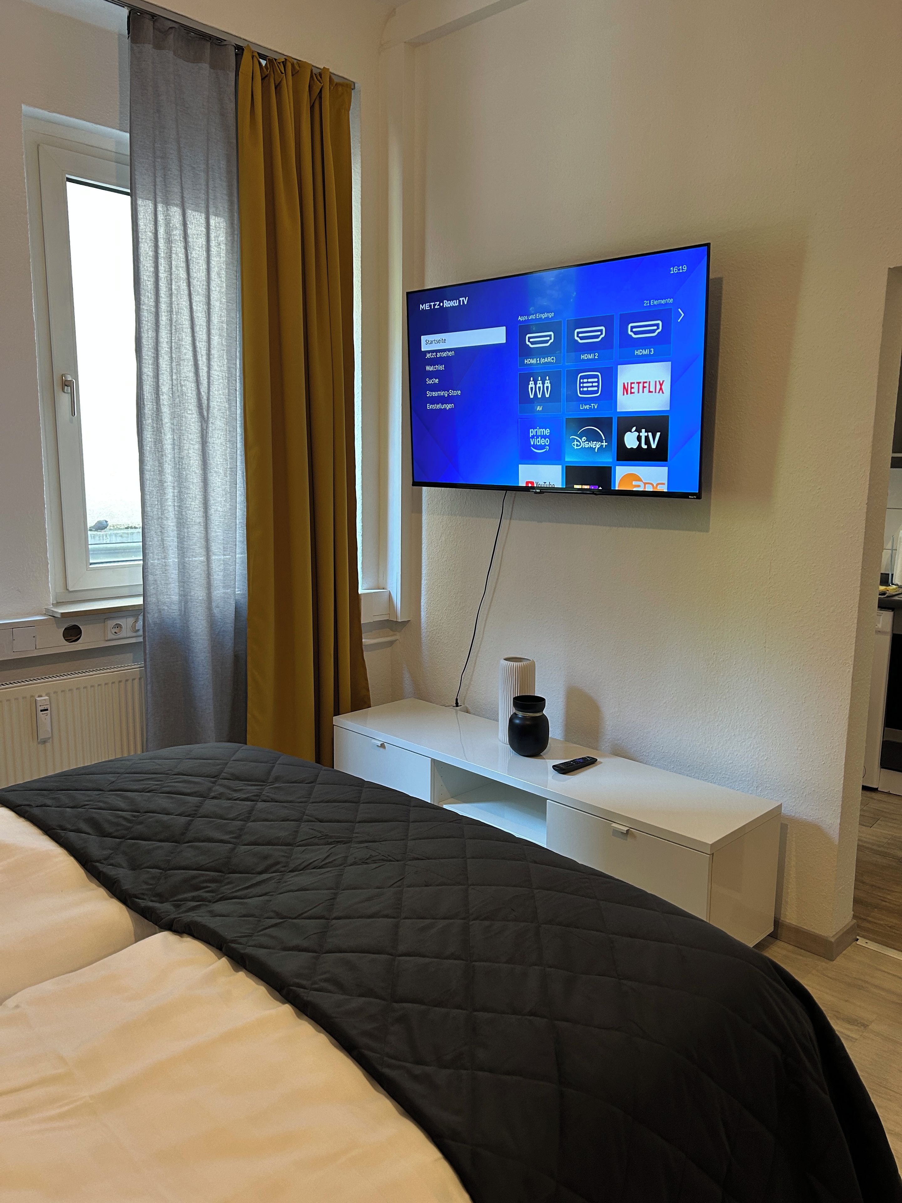 Ein Hotelzimmer mit einem Bett, einem an der Wand befestigten Fernseher, einem Nachttisch mit Fernbedienung und einem Fenster mit Vorhängen.