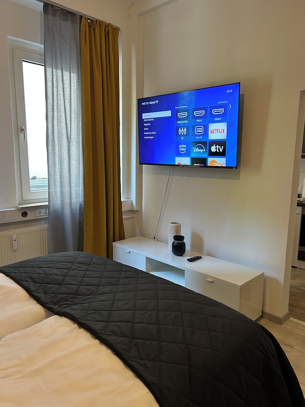 A hotel room with a bed, a TV mounted on the wall, a bedside table with a remote control, and a window with curtains.
