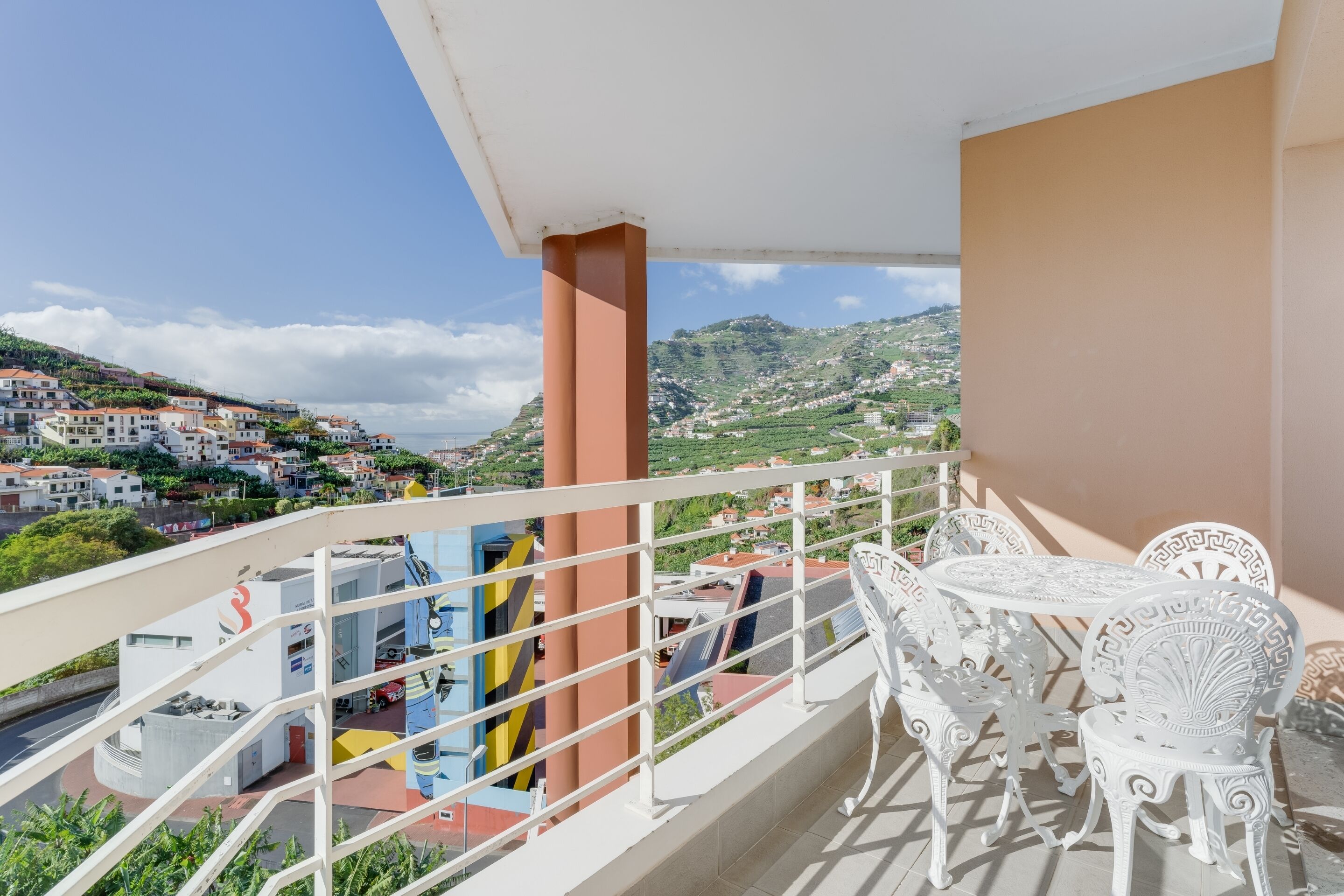 Un balcón con sillas y una mesa de metal blanco, con vistas a un pueblo en la ladera.