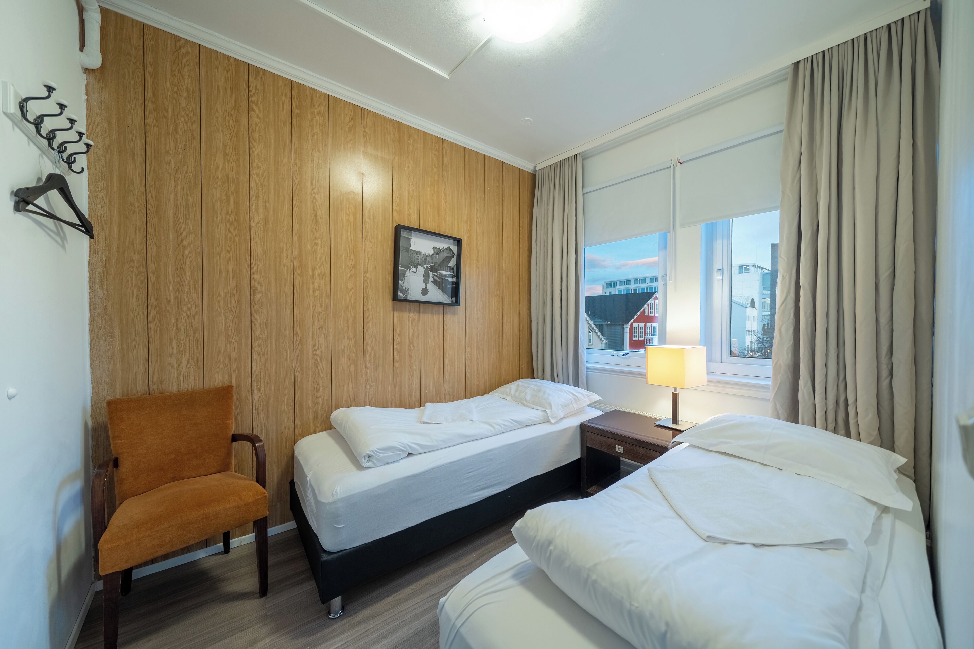 A hotel room with two beds, a wooden panel wall, a chair, a nightstand, a lamp, and a window with curtains.