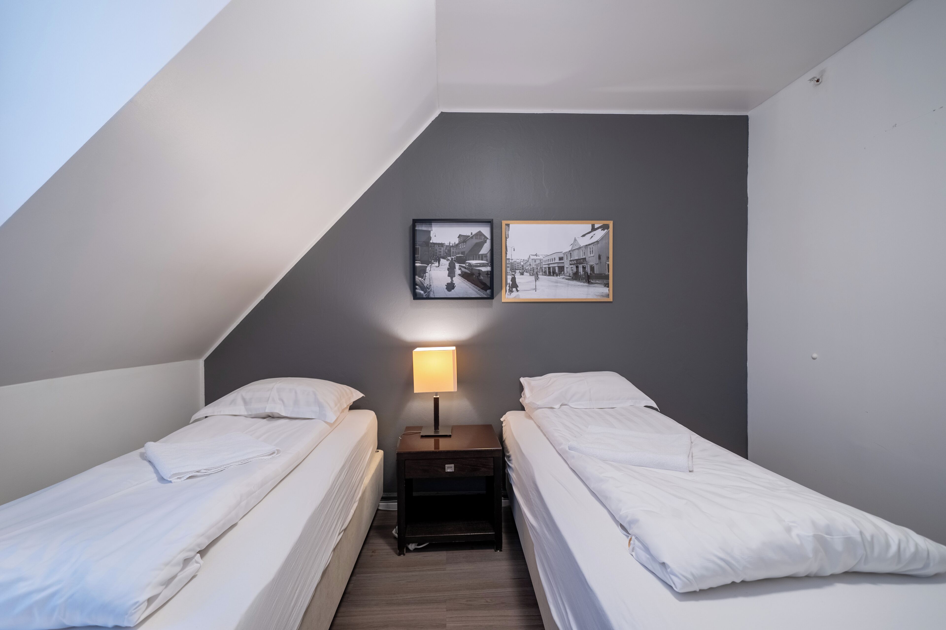 A small, modern room with two single beds, a nightstand with a lamp, and two framed pictures on the wall.