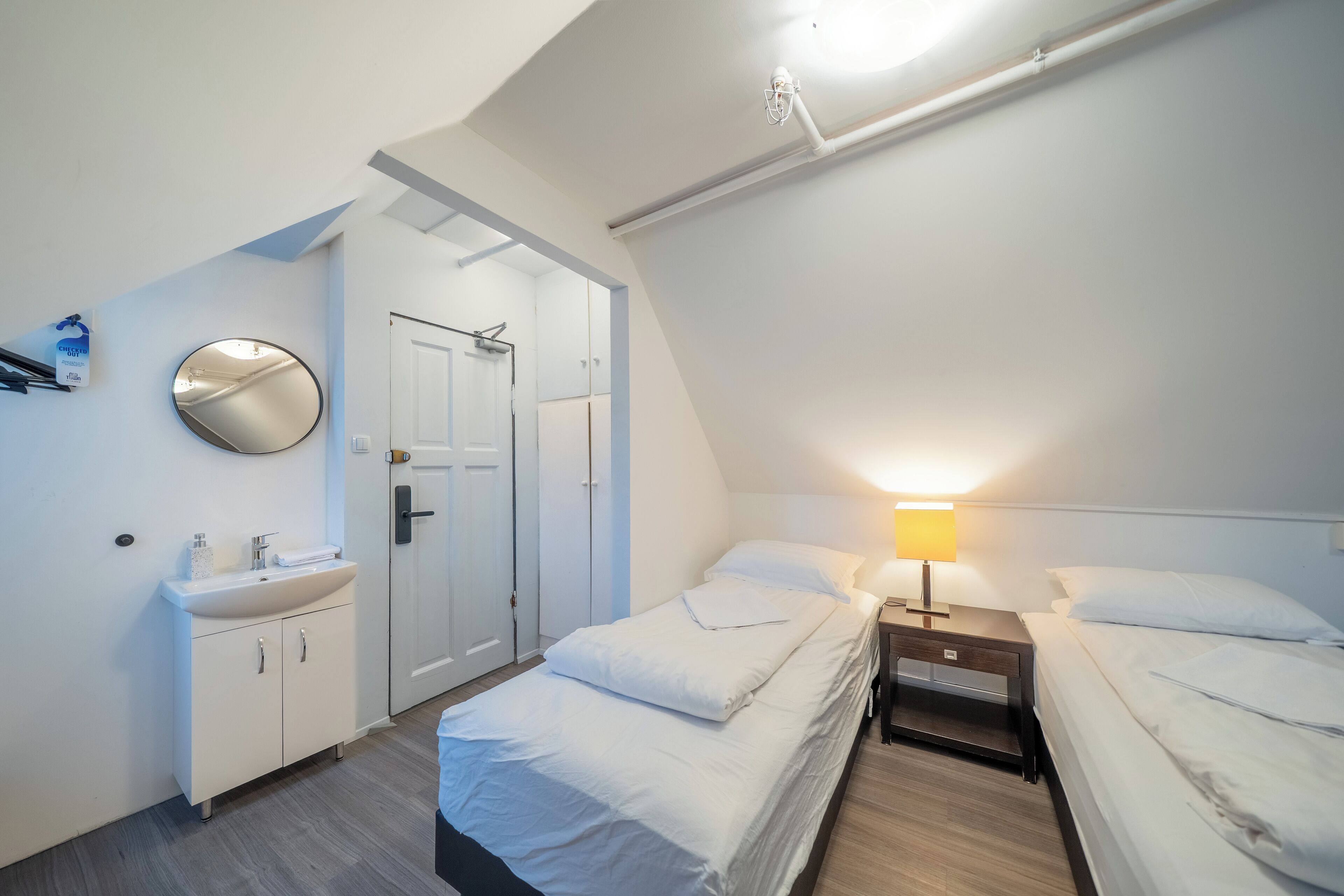 A bedroom with two beds, a bedside table with a lamp, a mirror, a white cabinet, and a door.