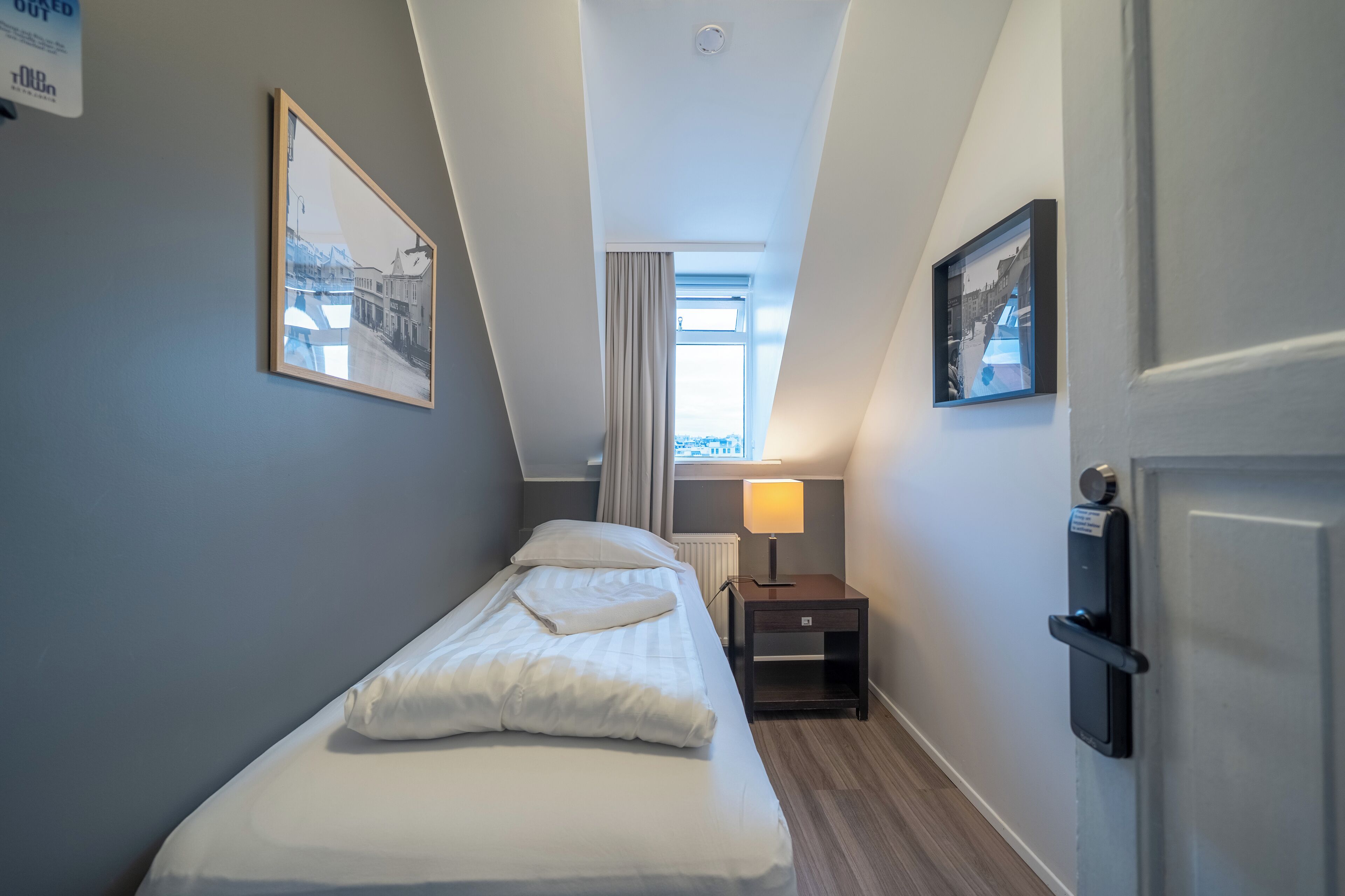 Habitación de hotel estrecha con una cama individual, una mesita de noche, una lámpara y una puerta con cerradura.