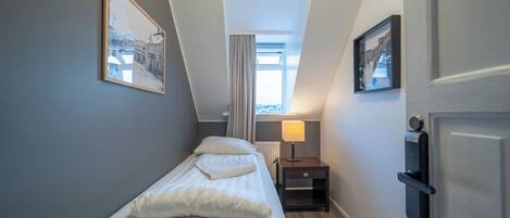 Habitación de hotel estrecha con una cama individual, una mesita de noche, una lámpara y una puerta con cerradura.