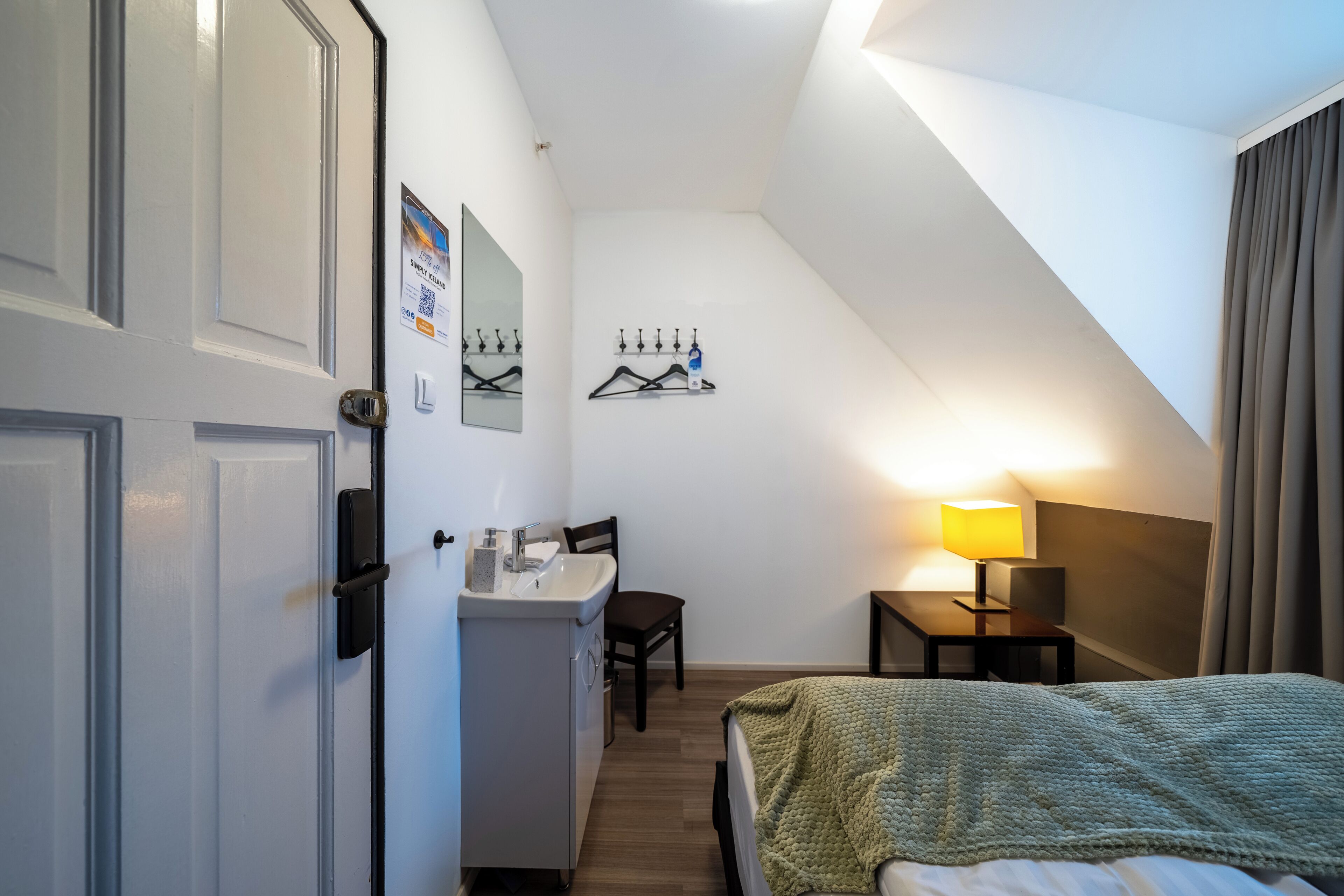 Een kleine hotelkamer met een bed, een bureau met stoel, een tafeltje met lamp en een badkamer met wastafel en spiegel.
