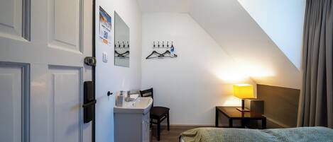 Habitación de hotel compacta con cama, escritorio con silla, pequeña mesa con lámpara y baño con lavamanos y espejo.