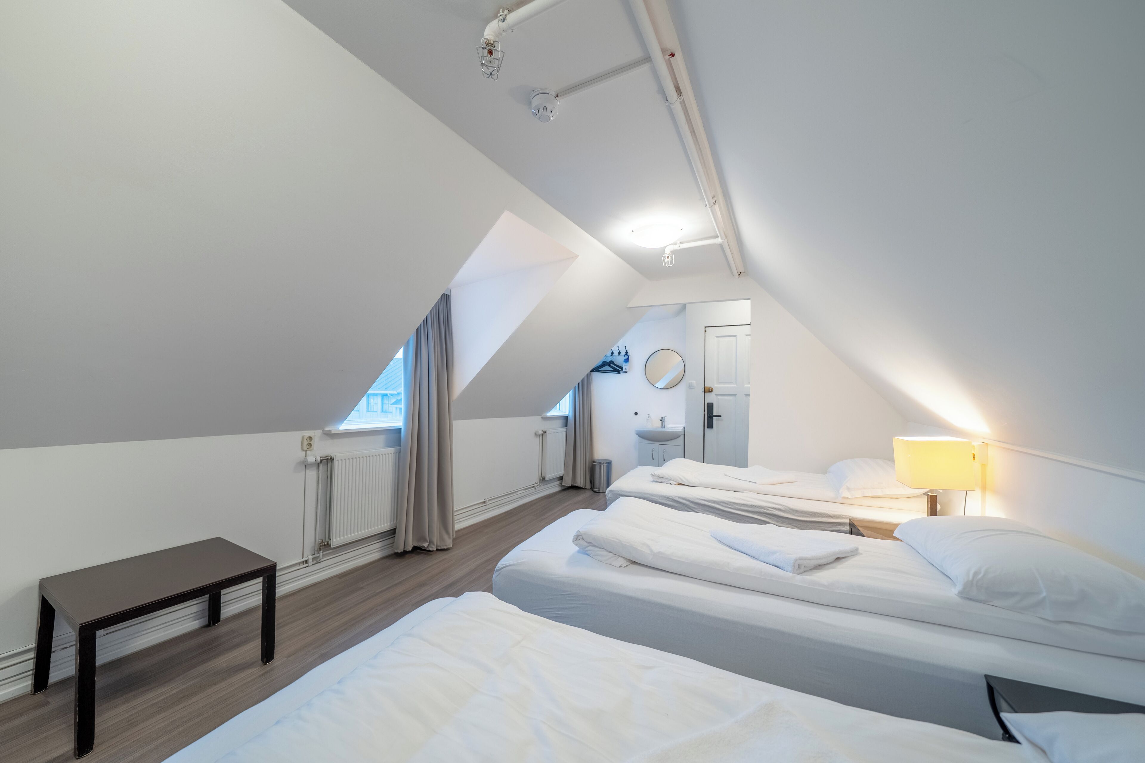 Une chambre moderne et bien éclairée, équipée de deux lits, d’une petite table et d’une salle de bain avec un lavabo et un miroir.