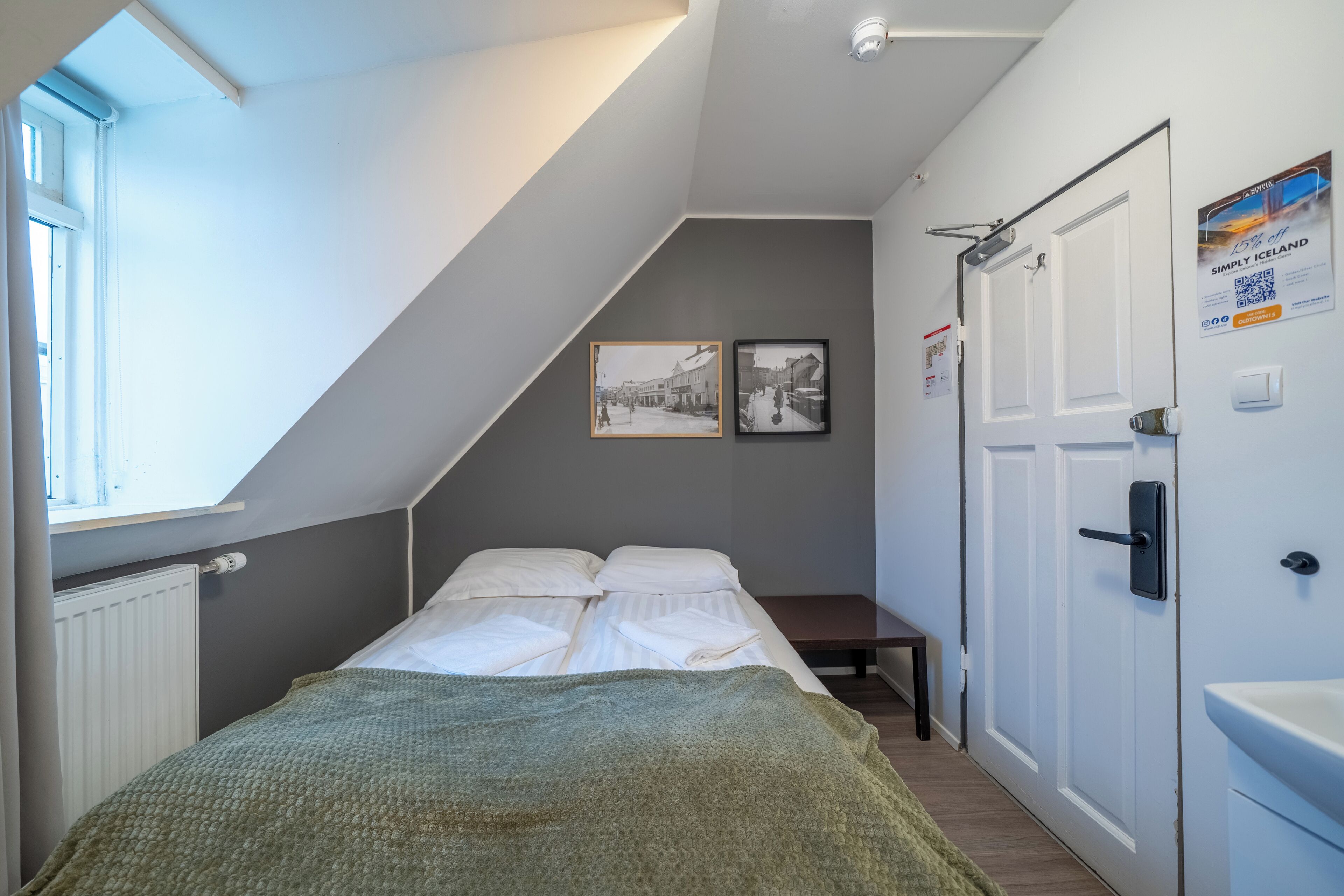 Un dormitorio con cama, ventana, radiador, puerta y dos cuadros en la pared.