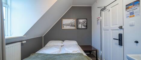 Un dormitorio con cama, ventana, radiador, puerta y dos cuadros en la pared.