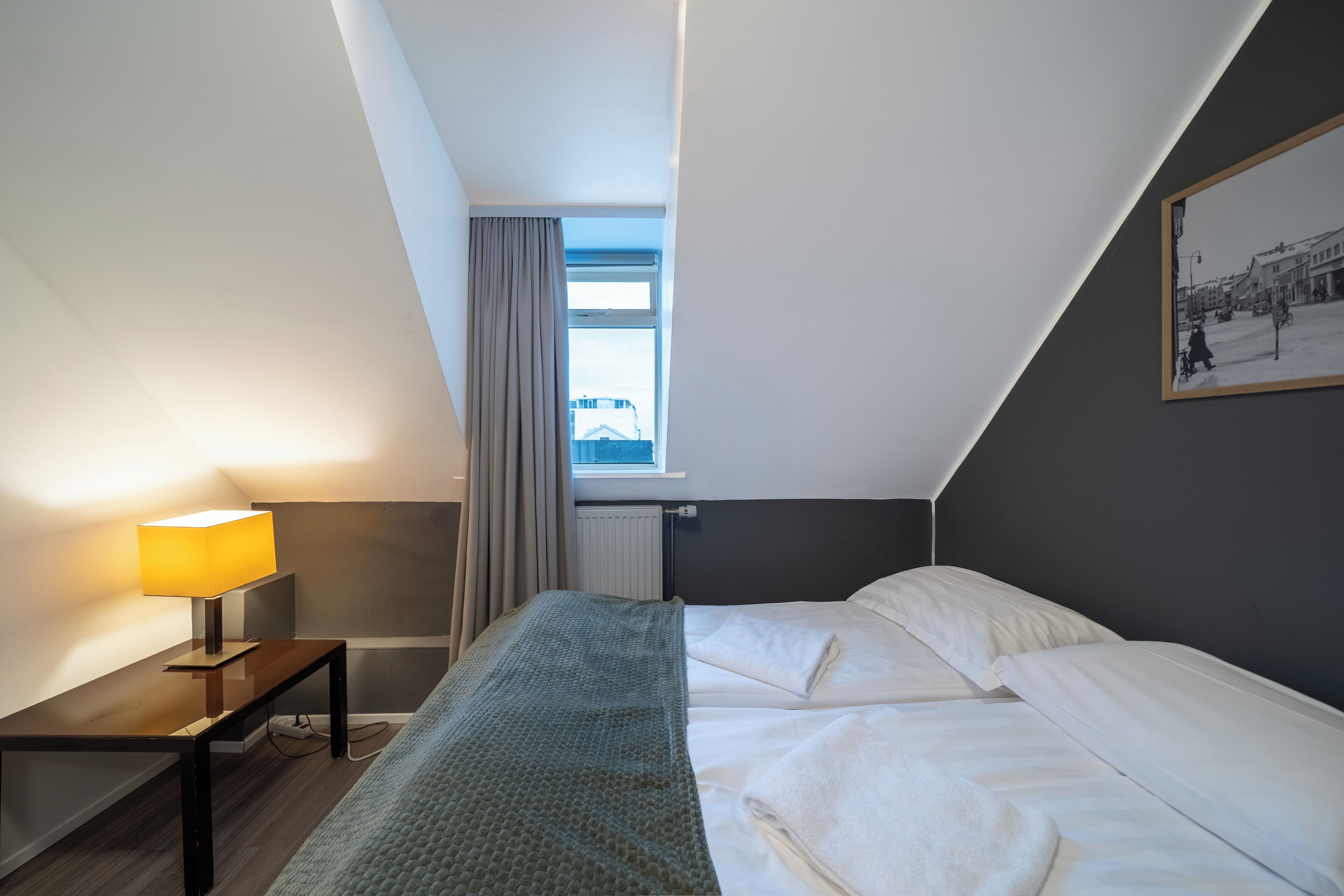 A modern hotel room with a bed, a bedside table with a lamp, a window with curtains, and a framed picture on the wall.