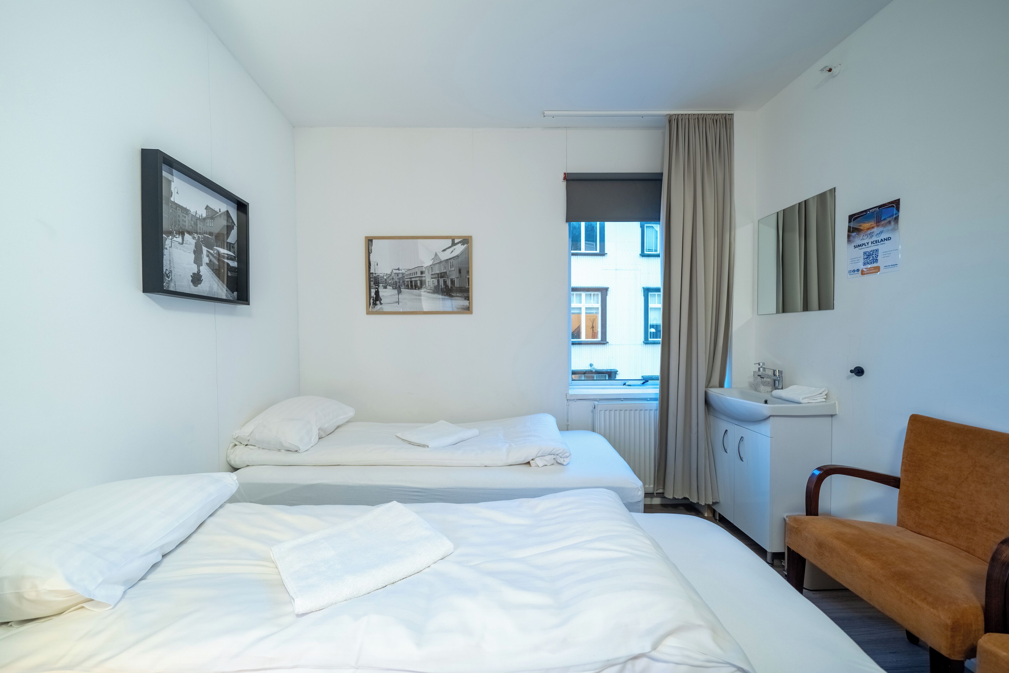 A hotel room with two beds, a small desk, a chair, and a window with a view of buildings.