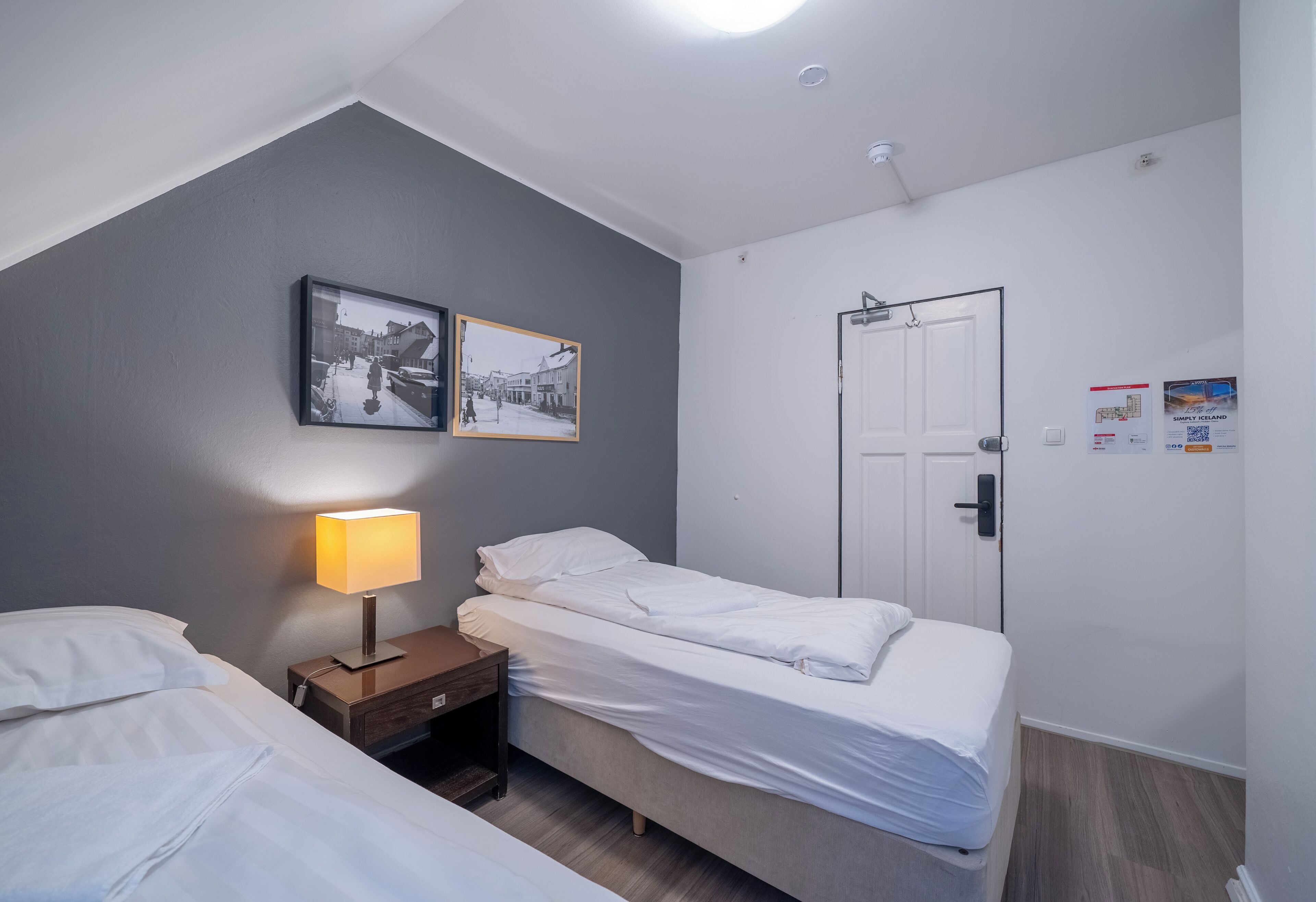 A small, modern bedroom with a single bed, a nightstand with a lamp, a door, and two framed pictures on the wall.
