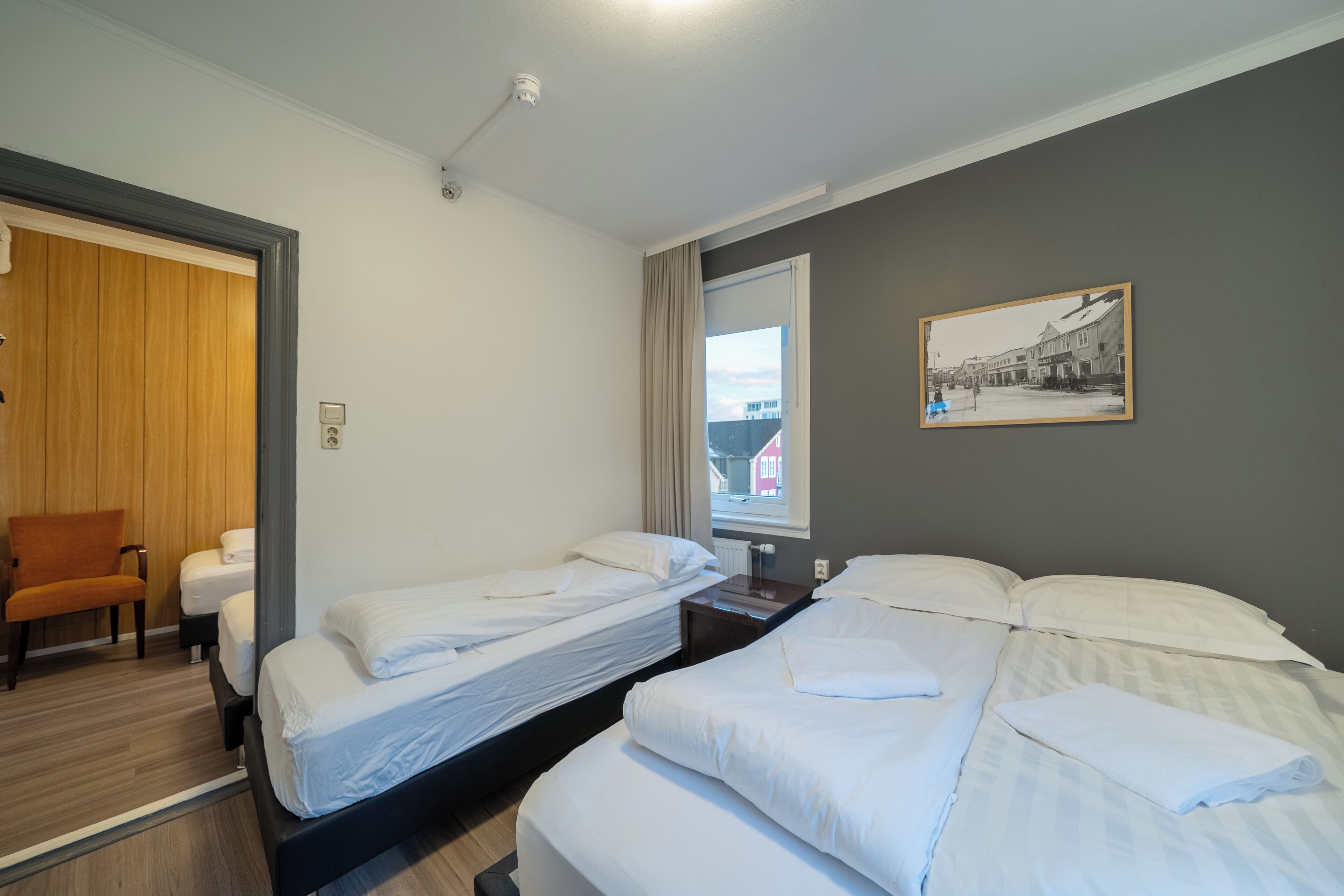 A hotel room with two single beds, a wooden chair, a small table, and a window with a view of buildings.