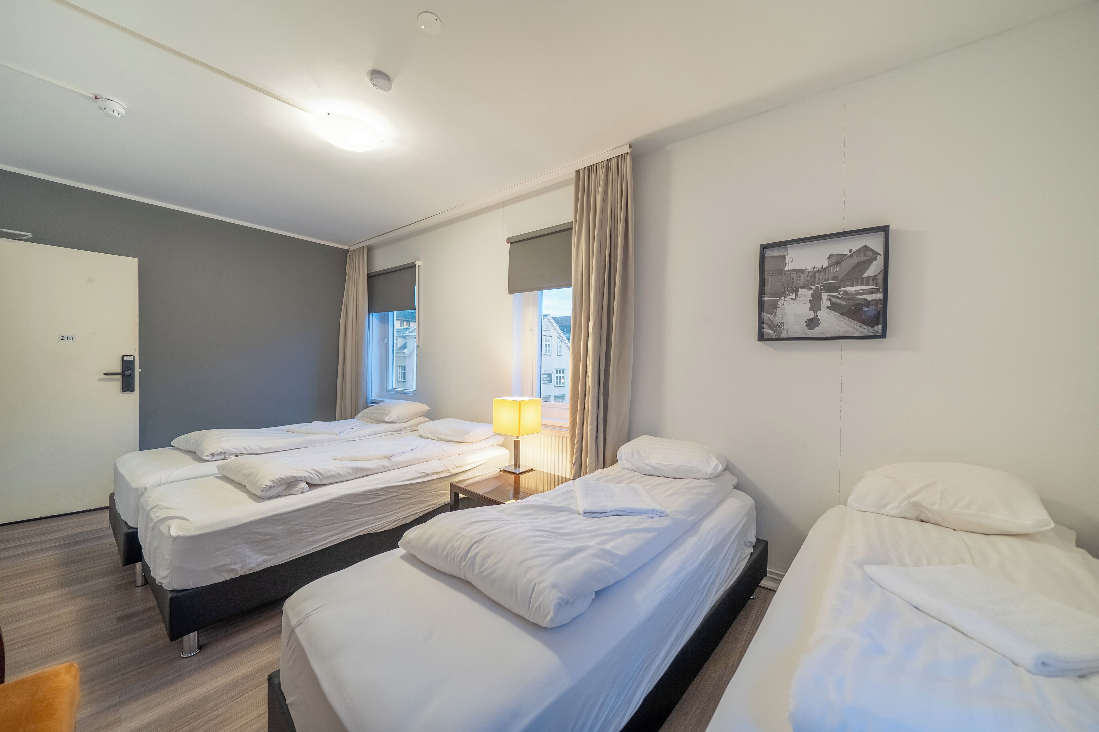A hotel room with three beds, a bedside table with a lamp, a framed picture on the wall, and a door with a number.