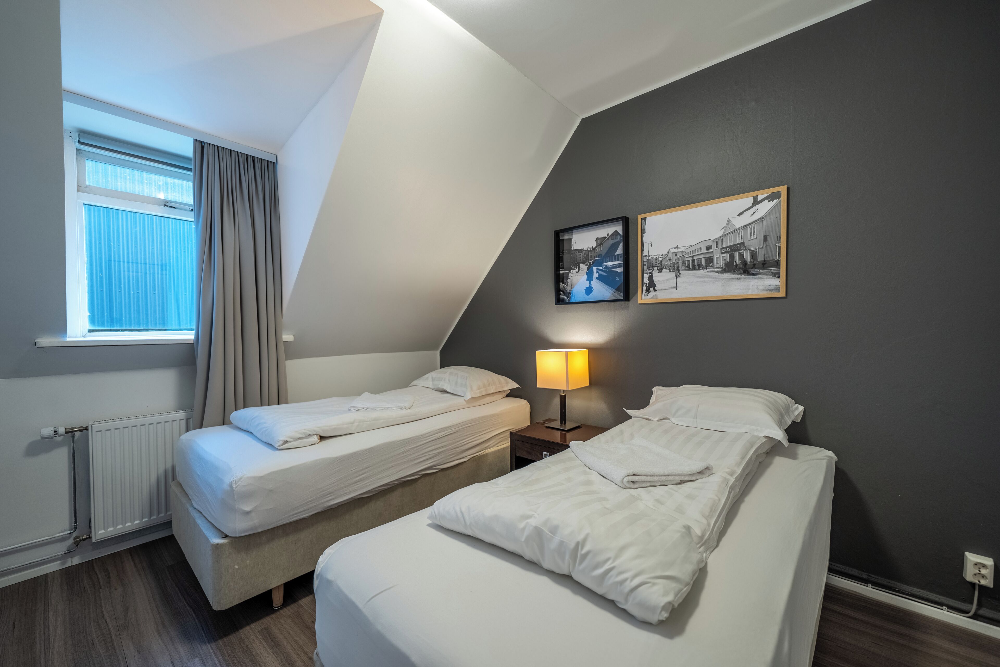 Ein Hotelzimmer mit zwei Betten, einem Fenster mit Vorhängen, einer Lampe und zwei gerahmten Bildern an der Wand.