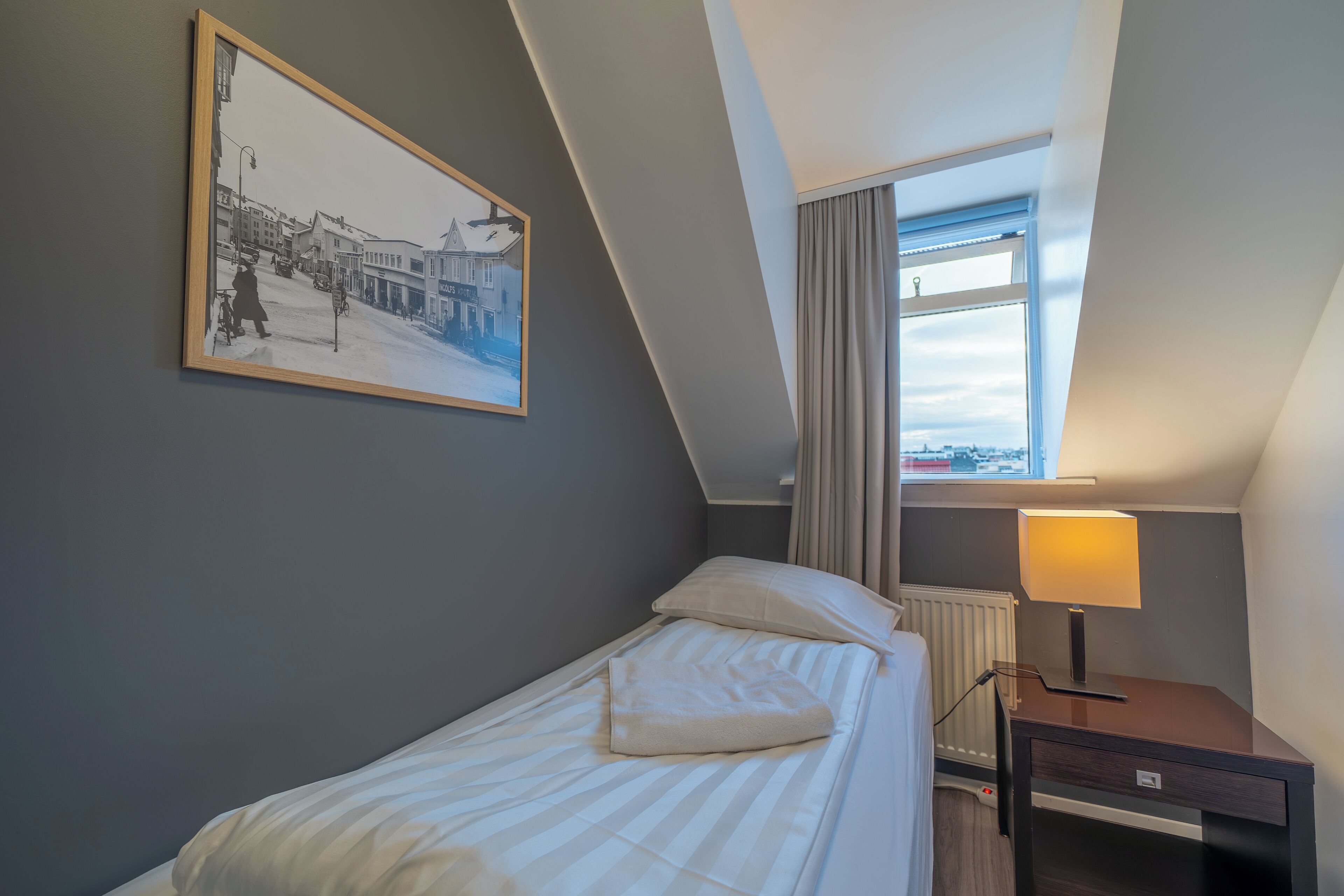 Una cama individual con almohada y mesita de noche con lámpara en una habitación con techo inclinado y ventana con cortinas.