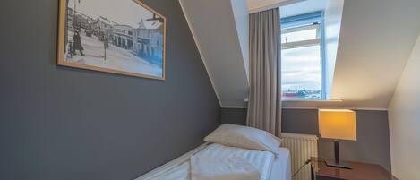 Una cama individual con almohada y mesita de noche con lámpara en una habitación con techo inclinado y ventana con cortinas.